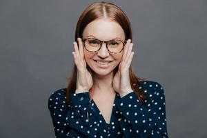 Photo of attractive woman with pleased facial expression, keeps both hands near cheeks, wears polka dot shirt, poses over grey studio wall, being happy. European pleased lady expresses happiness