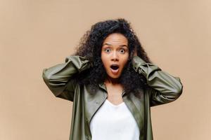 Impressed African American woman keeps both hands on head, opens mouth widely, dressed in white t shirt and leather shirt, being amazed by something, isolated over brown background. Omg concept photo