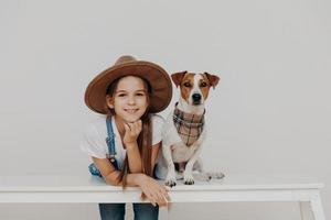 Content pretty girl wears hat, leans at white table, poses near pedigree dog, enjoy spending free time together, going to have walk, rest at home. Children, happiness, animals, lifestyle concept photo