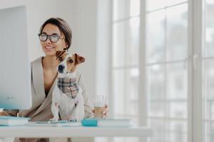 Photo of woman works freelance from home, concentrated in monitor of computer, wears spectacles, poses at desktop with jack russel terrier dog, drinks beverage, smiles positively enjoys favorite work