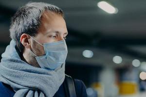 Pensive man wears protective mask against new coronavirus from China, wrapped scarf around neck, looks somewhere, thinks about epidemic situation. Influenza, flu symptoms, virus, treatment concept photo