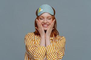 Pleased woman touches cheeks, has healthy freckled skin, smiles broadly prepares for sleep, wears striped pajama and sleep mask, closes eyes, isolated over grey background. Rest and lifestyle concept photo