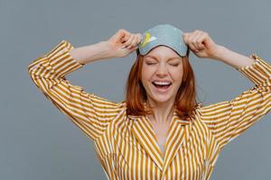 Overjoyed red haired Caucasian woman laughs positively, wears blindfold and yellow striped pajama, expresses good emotions, isolated on grey background. Good rest, bedtime and sleeping concept photo