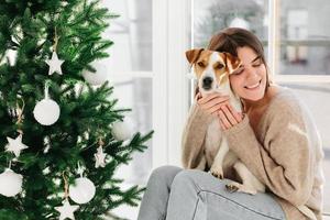 Horizontal shot of caring smiling woman hugs puppy with love, enjoys cozy atmopshere and lovely moment, smiles pleasantly, decorate New Year tree together, celebrate favourite holiday at home