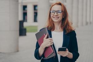 Happy red-haired business lady in official clothes standing outdoors, using mobile phone photo