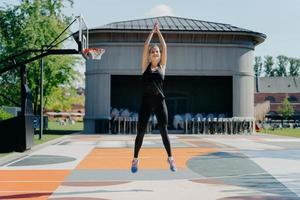 Athletic young woman in black activewear jumps high keeps arms raised up clasps hands warms up before jogging has training on stadium outdoors has cardio burns fat and calories for weight loss photo