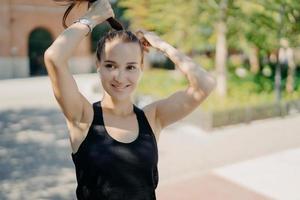 Positive sportswoman combes pony tail being in good physical shape listens music in earphones wears casual t shirt looks somewhere poses outdoor enjoys good weather sunny day. Sport concept. photo