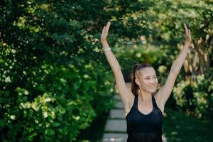 Photo of cheerful young woman with smile on face keeps arms raised up being in good mood dressed in active wear poses against green nature background. People healthy lifestyle recreation concept