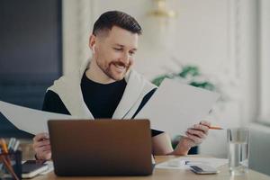 Handsome happy businessman analyzing documents and feeling satisfied while working on laptop from home photo