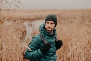 Shot of handsome young man with dark stubble, carries backpack, looks back, notices something, has thoughtful expression, spends free time in open air. Autumn time. People and adventure concept photo