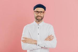 Indoor shot of self confident unshaven man looks happily at camera, keeps arms folded, wears headgear and shirt, listens attentively interlocutor, isolated over pink background. Masculinity concept photo