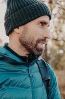 Sideways shot of unshaven man with dark thick beard, wears warm har and anorak, looks pensively aside, poses outside, dreams about something has outdoor stroll in unknown place alone, active lifestyle photo