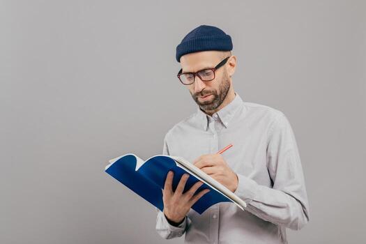 Serious male teacher writes down notes for next class, holds notepad, prepares interesting task for students, wears formal outfit, stands against grey background with copy space on right side for text photo