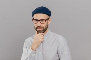 Blue eyed male adult holds chin and looks attentively at camera, ponders on way out or solution, wears spectacles, headgear and formal shirt, models over grey studio wall. Businessman poses indoor photo