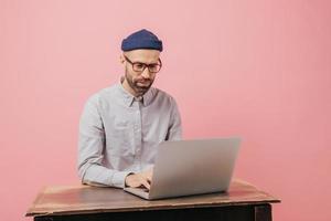 Concentrated hipster student reads information and checks data, focused in monitor of laptop computer, keyboards something, sits at desk, wears formal clothes, isolated over pink studio wall photo