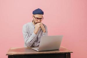 Horizontal shot of concentrated male worker reads statistics attentively, focused in laptop computer, learns something online, wears spectacles, hat and white shirt, isolated over pink wall. photo