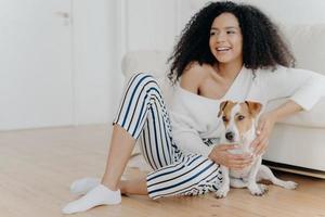Cute cheerful young African American female sits on floor near sofa, plays with dog, dressed in stylish clothing, looks gladfully somewhere, spends free time at home. Happy pet owner indoor. photo