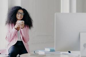 Joyful curly haired female freelancer or blogger has coffee break, holds white mug with beverage, poses near window, focused in computer, watches useful training video, wears near formal clothes photo