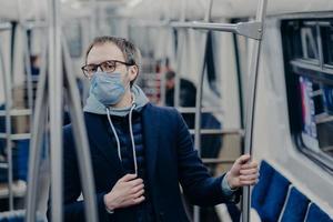 Serious European man being aware of his health, protects from catching serious disease in public transport, wears medical mask on face, travels in urban train, afraids of coronavirus epidemy photo