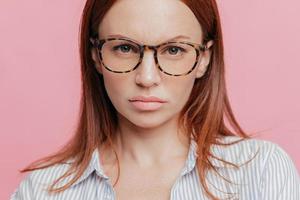 Close up shot of lovely female model wears big optical glasses, has brown hair, looks seriously with confident expression, isolated over pink background, thinks about business affairs. Headshot photo