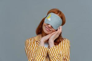 Pretty redhead woman wears blindfold on eyes, keeps hands pressed together near face, sees pleasant dreams while sleeping, dressed in domestic clothes, isolated on grey wall, feels comfortable photo