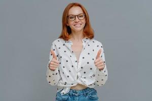 Positive redhaired woman with gentle smiles shows thumbs up, demonstrates like or approval gesture, says well done, likes someting, wears polka dot shirt and jeans, isolated on grey studio wall photo