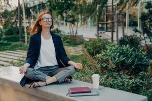 Peaceful business woman holding hands in mudra gesture, sitting in lotus pose and meditating in park photo