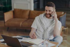 Cheerful man freelancer makes telephone call writes down information in notepad makes notes in planner looks attentively at laptop screen poses against home interior at desktop. Technology user photo