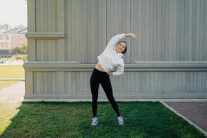 Sporty lifestyle wellness and weight loss concept. Active woman in sportwear trains outdoors leans left stands on green grass warms up before workout has active life enjoys regular workout in open air photo