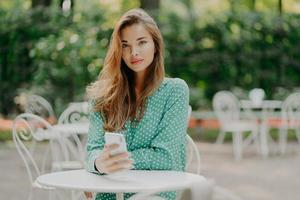 Outdoor shot of fashionable woman with long hair, dressed in green polkadot shirt, uses modern cellphone for communication, connected to internet, sits at white table in outside cafe, waits for order