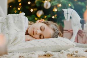 Close up shot of attractive small child lies on floor, sleeps near New Year or Christmas tree, warms with white knitted sweater, has pleasant dreams in evening. Children, winter holidays, rest concept photo