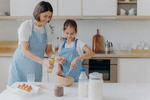 Brunette mother adds oil in dough, little daughter helps to make pastry, whisks ingredients, pose together against kitchen interior, prepare bakery together. Small helper with mommy. Homemade food photo