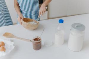 Cropped view of kid in apron whisks ingredients in bowl with beater, busy cooking and helping mother to prepare cake. Dough, milk, eggs, wooden spatula and chocolate on table photo