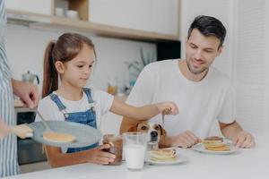 foto de una niña con peto de mezclilla agrega chocolate a los panqueques, desayuna junto con papá y el perro, le gusta cómo cocina la madre. la familia en la cocina desayuna durante el fin de semana. momento feliz