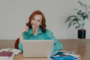 Beautiful redhead woman works distantly from home, sits in front of laptop computer finishes working on project thinks about adding interesting content on own website prepares publication for web page photo