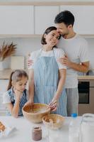 Family cook meal at home. Husband and wife embrace with love, small girl poses near, prepare delicious dinner, use milk, chocolate, eggs, make homemade pastry, pose against kitchen interior. photo