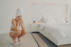 Young sad beautiful woman in white underwear standing on electronic smart scales in bedroom photo