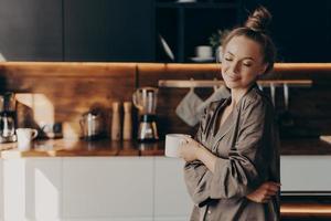 Beautiful young happy woman standing in cozy silk pajamas in kitchen with cup of coffee photo