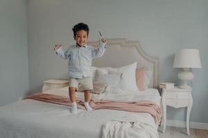 alegre pequeño niño afroamericano saltando en la cama en casa y sonriendo foto