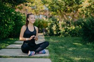 Cheerful female runner poses in lotus pose outdoor dressed in sportswear uses mobile phone during rest after jog downloads music songs in player prepares for morning workout chooses audio records