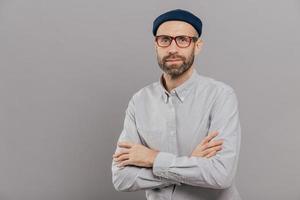 Photo of serious male keeps arms folded over chest, dressed in white formal shirt, fashionable hat, poses for making photography over grey wall with free space for your advertisement or promotion