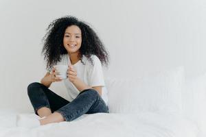 Young pretty African American woman rests in bed at home, drinks hot tea in morning, enjoys domestic atmosphere, tasty drink while relaxes in bedroom. People, leisure, rest and lifestyle concept photo