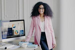 Half length shot of cheerful African American woman with curly hair, dressed in formal elegant outfit, poses near table, computer screen with graphics, prepares for presentation in front of auditorium photo