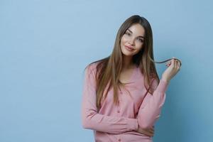 Waist up shot of pleasant looking young woman has beautiful long hair, tilts head, dressed in casual jumper, has perfect clean skin, advertises shampoo for hair care, isolated on blue background. photo