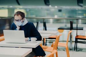 Man freelancer wears face mask to prevent spread of coronavirus and coughing, uses laptop computer for distant work, poses in cafe with cup of coffee, reads information about symptoms of disease photo