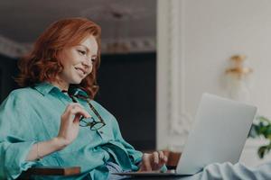 Side view of pretty redhead woman concentrated on remote job, prepares publication for web page, looks gladfully at laptop computer, sits at modern apartment, works remotely, holds eyeglasses photo