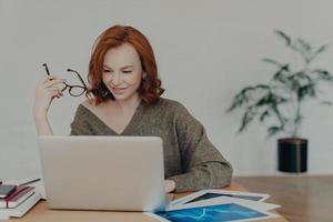 Busy redhead adult woman does freelance work on laptop computer, focused in monitor, prepares project, surrounded with paper documents, wears warm sweater, analyzes information, uses internet photo