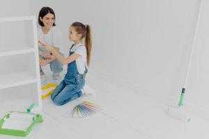 Busy girl and her smiling mother paint drawer in white color, use paintbrush or roller, surrounded with color samples, pose in white empty room, think about house decoration. Domestic work concept photo