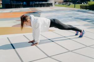 Full length shot of active young woman with pony tail stands in plank focused down dressed in active wear poses outdoors being motivated gets stronger demonstrated endurance and determination photo