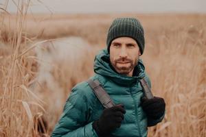 Photo of attractive man with contemplative expression, wears gloves, hat, poses outdoor, looks aside, walks across field, thinks about something. Cold weather. Masculinity concept. Lifestyle